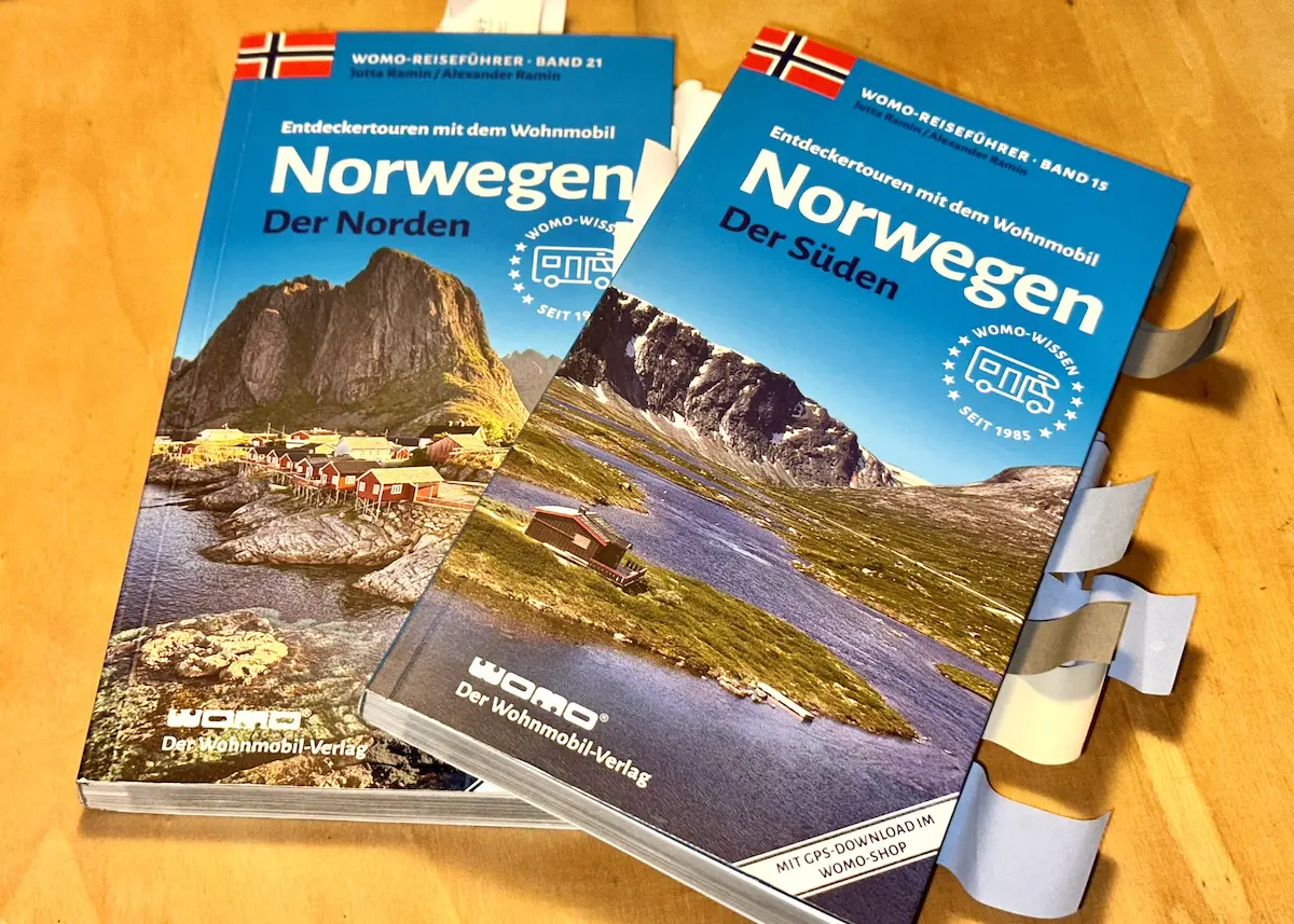 Auf einem Tisch liegen zwei Reiseführer für Norwegen: der eine mit dem Titel „Norwegen – Der Norden“, der andere „Norwegen – Der Süden“ – perfekte Begleiter, wenn Sie eine Reise nach Norwegen planen. Im Reiseführer für den Süden sind Seiten mit Haftnotizen markiert, was auf eine sorgfältige Vorbereitung hindeutet. Beide Einbände zeigen malerische Landschaften, die den Reiz norwegischer Abenteuer einfangen.