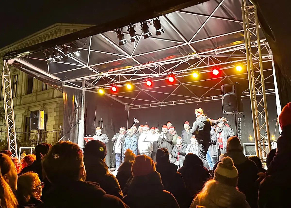Eine Gruppe von Menschen mit Weihnachtsmützen tritt auf einer Freiluftbühne auf einem alternativen Berliner Weihnachtsmarkt auf, beleuchtet von Scheinwerfern und von einer Menschenmenge aus betrachtet.