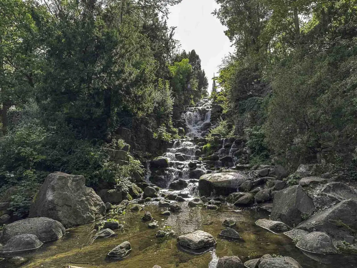 Ein kleiner Wasserfall stürzt in Kaskaden über Felsen, umgeben von üppig grünen Bäumen und Laub, mit einem flachen Bach und großen Steinen im Vordergrund, die bei klarem Himmel an malerische Orte in den Berliner Parks erinnern.
