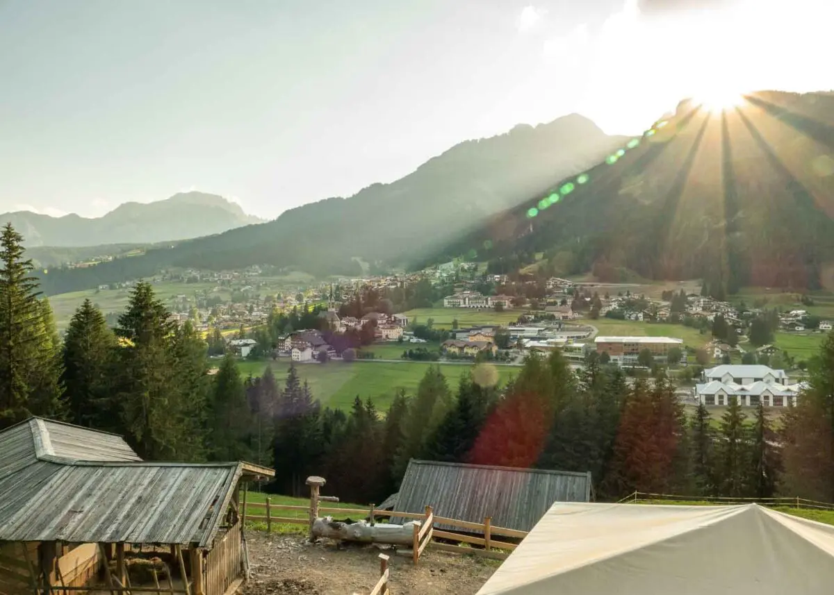 Blick über eine Almhütte ins Fassatal, während die Sonne an einem Gipfel der Dolomiten untergeht.