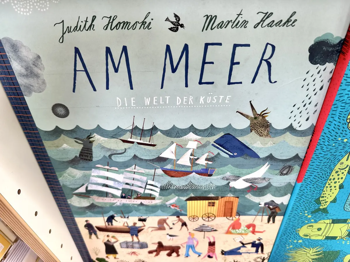 Das Cover des Kinderbuchs mit dem Titel „Am Meer“ zeigt illustrierte Schiffe, Wellen, Strandbesucher, Meeresbewohner und Tiere. Die Namen der Autoren, Judith Homoki und Martin Haake, sind oben zu sehen, darunter steht „Die Welt der Küste“. Dieses farbenfrohe Cover eignet sich perfekt für die Reihe „Kinderbücher Reise“ und lädt junge Leser dazu ein, Abenteuer an der Küste zu erleben.