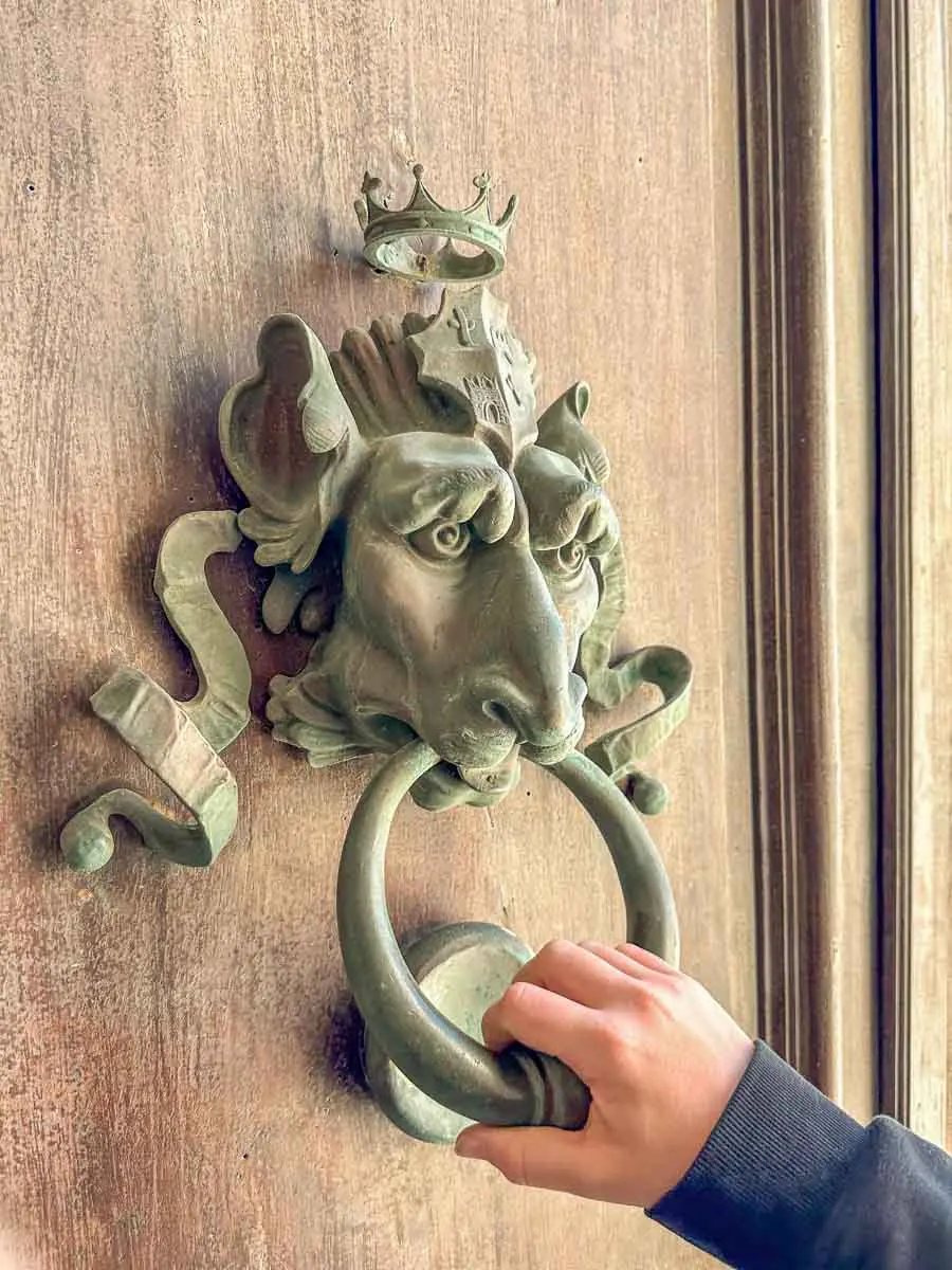 Eine Kinderhand bewegt einen kunstvoll geschmiedeten Türknauf, der einen Kopf mit Eselsohren und langer Nase zeigt an einer massiven Holztüre.