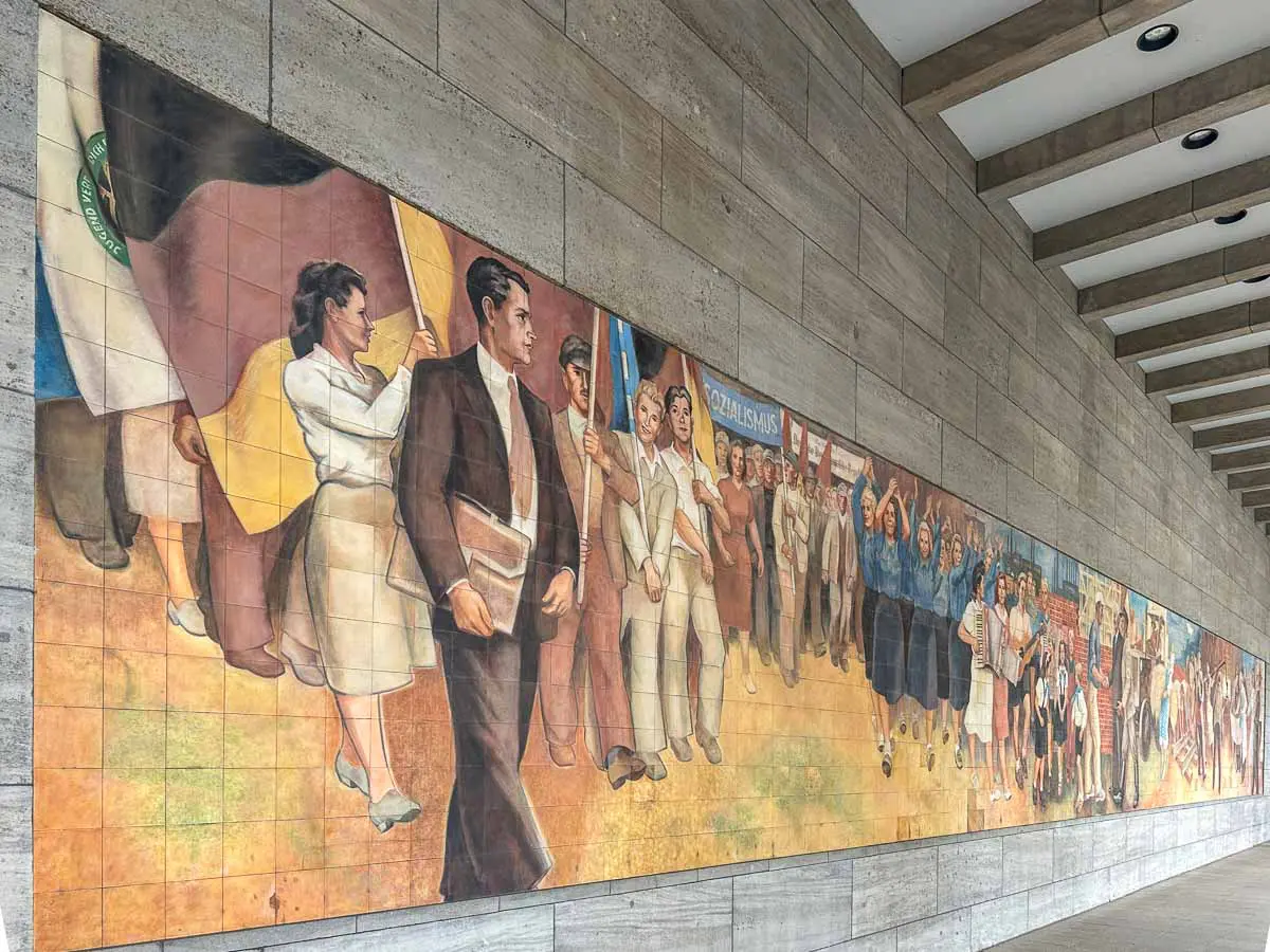 Ein propagandistisches Wandgemälde aus der DDR an einer gekachelten Wand in Berlin zeigt Menschen, die marschieren, Fahnen und Transparente tragen und unterschiedliche Kleidung tragen – ein Symbol für Zusammenhalt und Engagement. Das Wandgemälde erstreckt sich untr Arkaden mit rechteckigen Säulen.