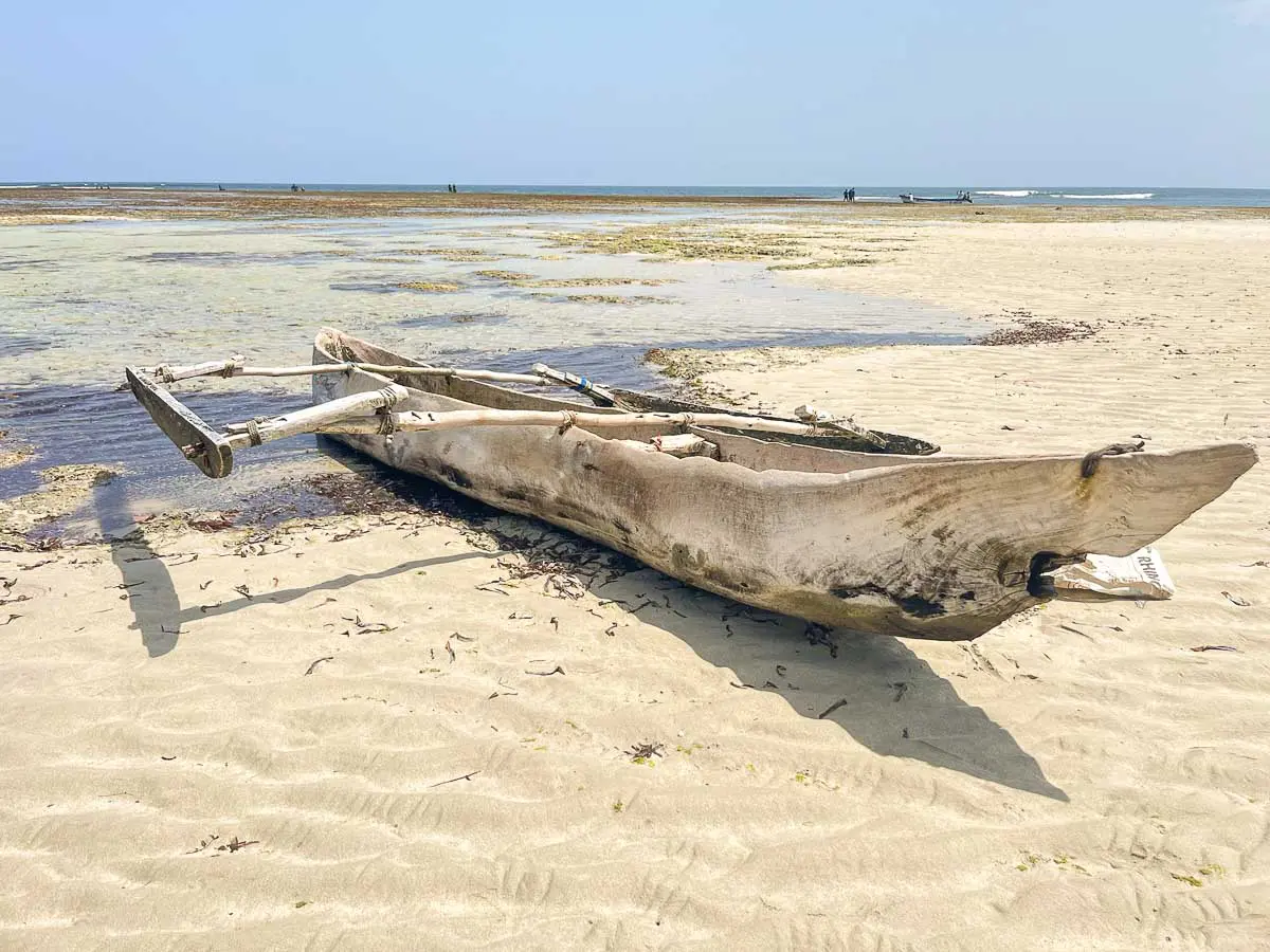 Ein verwittertes hölzernes Auslegerkanu liegt auf einem Sandstrand in der Nähe der Küste am seichten Wasser und das Meer unter einem klaren Himmel.