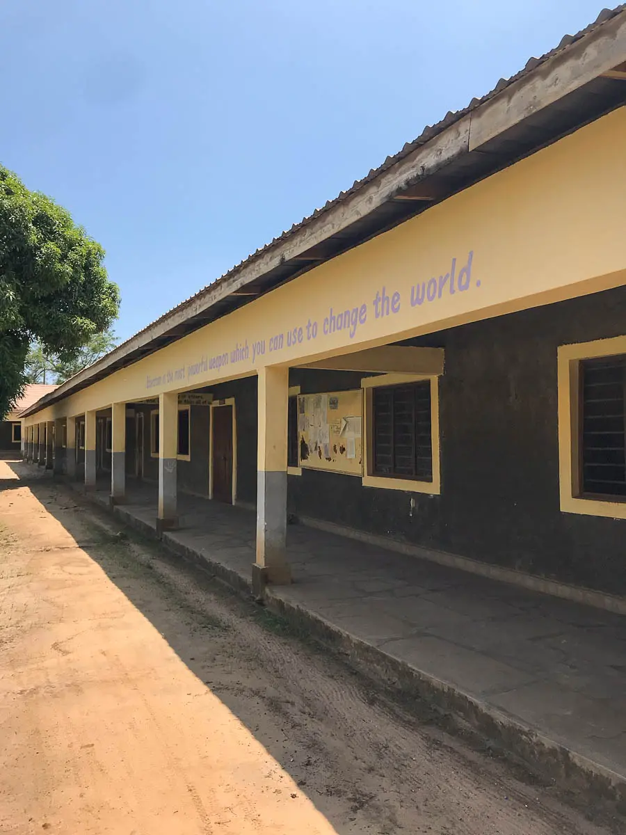 Ein langes, einstöckiges Schulgebäude mit gelben Verzierungen und einem schattigen Gang. Ein Zitat über dem Gehweg lautet: "Bildung ist die mächtigste Waffe...". Bäume und blauer Himmel vervollständigen diese Szene aus Kenia.