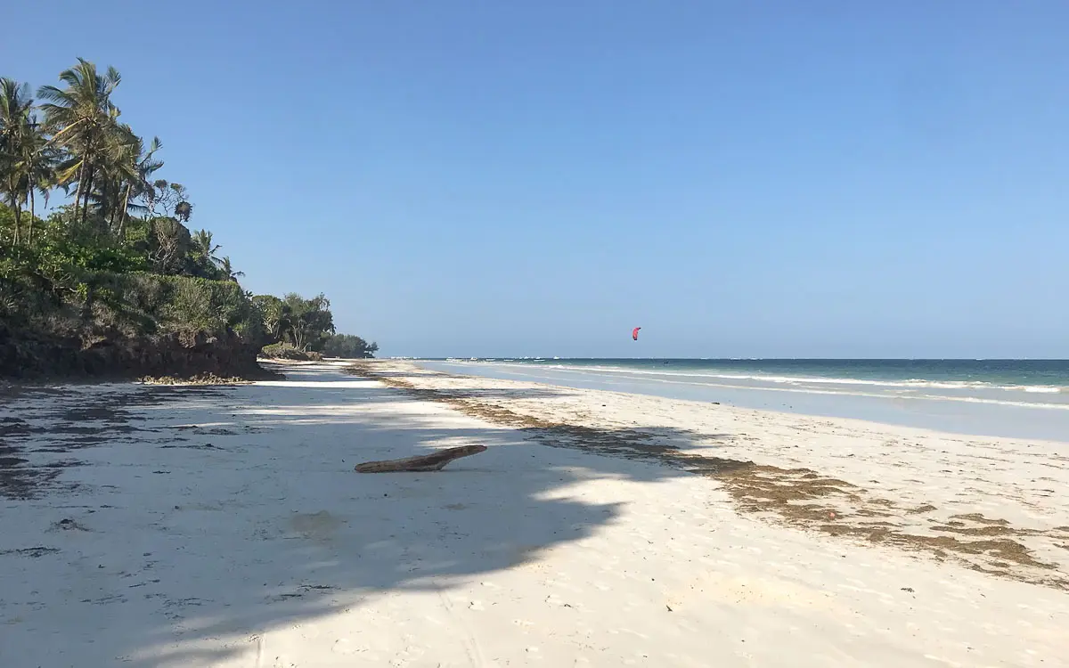 Ein sonniger, leerer Strand mit weißem Sand, Palmen auf der linken Seite und sanften Wellen - perfekt für eine Reise nach Kenia mit Kindern. Schatten erstrecken sich über den Sand, während in der Ferne ein einzelner roter Drachensurfer über das blaue Meer gleitet.