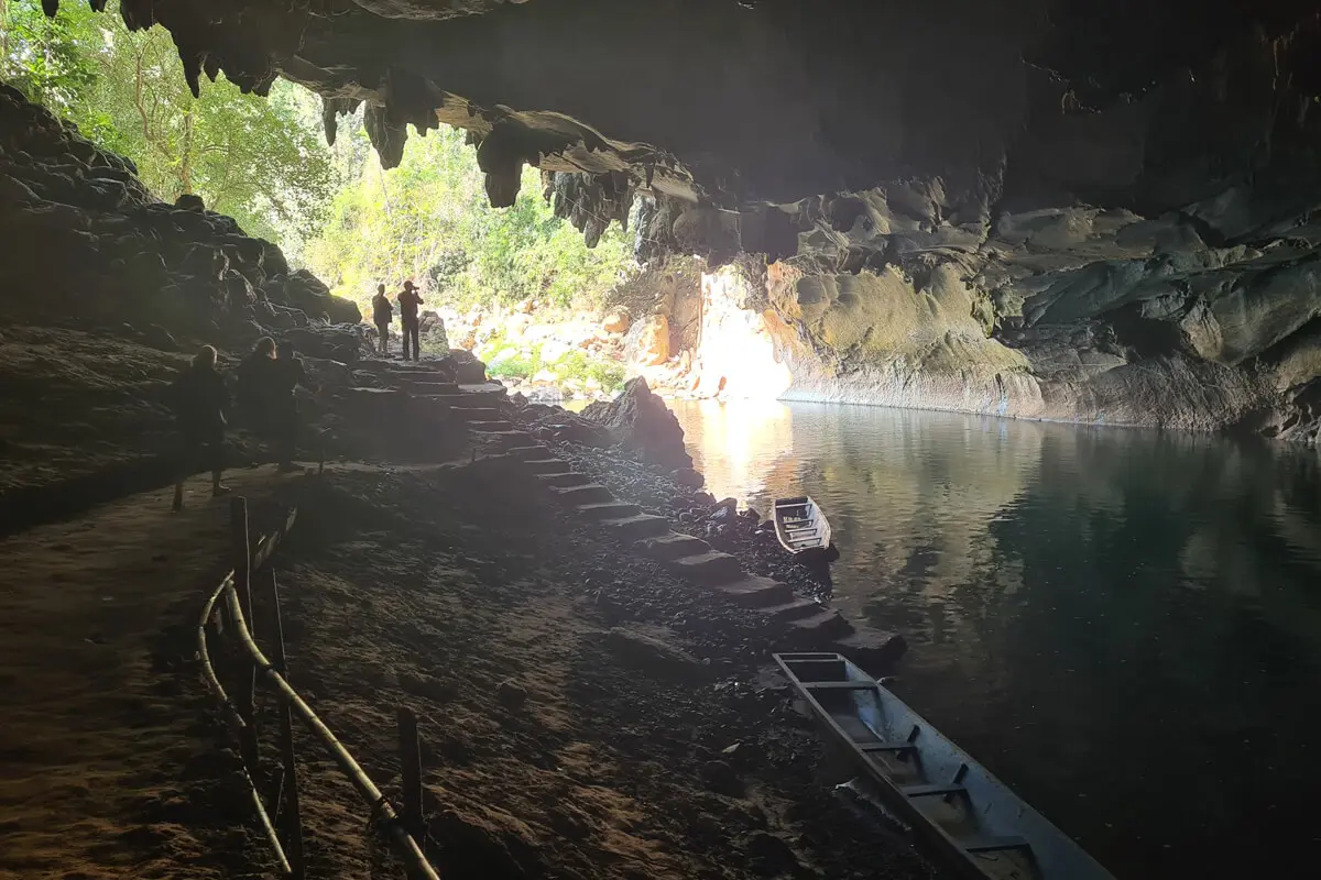 Silhouettierte Menschen, vielleicht eine Familie auf Abenteuerurlaub mit Kindern, gehen einen felsigen Pfad entlang, der in eine Höhle mit einem ruhigen Fluss führt; Licht strömt durch den üppig begrünten Eingang herein, und kleine Boote ruhen am Rande des Wassers.