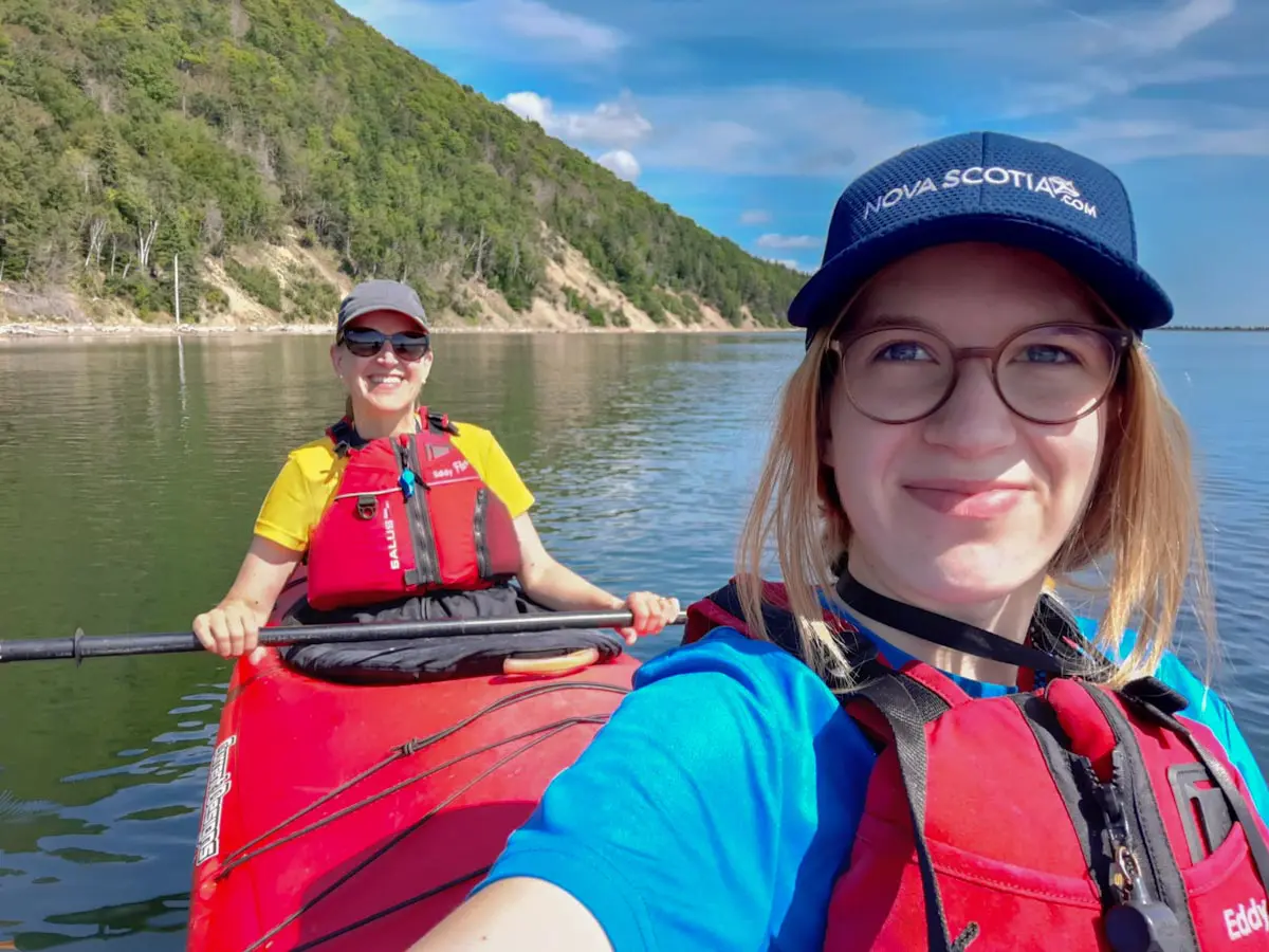 Mutter und Tochter in Schwimmwesten lächeln beim Kajakfahren auf ruhigem Wasser in der Nähe eines bewaldeten Ufers unter blauem Himmel. Die eine hält ein Paddel in der Hand, die andere, mit einem Nova Scotia Hut, macht ein Selfie - und hält damit ihr unvergessliches Reiseabenteuer fest.