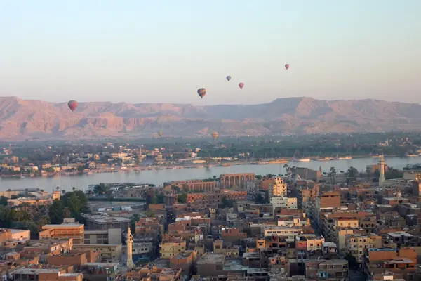 Wir schauen aus einem Heißluftballon auf Luxor und den Nil: eine Stadtlandschaft mit hellbraunen Gebäuden im Vordergrund, einem Fluss in der Mitte und Bergen im Hintergrund. Über dem Fluss schweben Heißluftballons - ein unvergessliches Reiseabenteuer.