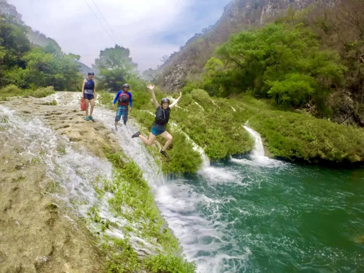 Drei Menschen stehen auf einem Felsen am Wasserfall, einer springt ins türkisfarbene Wasser. Üppiges Grün umgibt sie, Hügel steigen im Hintergrund auf. Perfekt für ein Reiseabenteuer oder einen Abenteuerurlaub mit Kindern und Familie.