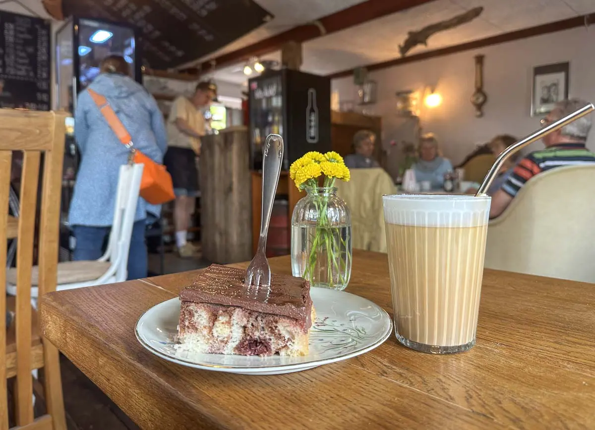Ein Stück Kuchen mit einer Gabel und ein Glas Kaffee mit einem Metallstrohhalm stehen auf einem Holztisch in einem gemütlichen Café - perfekt zum Entspannen nach der Erkundung von Campingplätzen oder Wohnmobil-Stellplätzen in Strandnähe. Gelbe Blumen erhellen den warm beleuchteten Hintergrund.