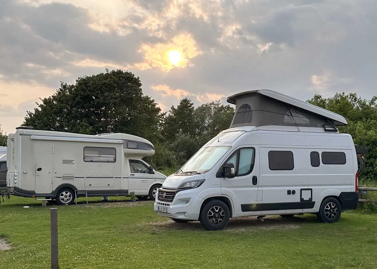 Zwei weiße Wohnmobile stehen auf einem Campingplatz an der Ostsee auf einer Wiese vor Bäumen unter einem bewölkten Sonnenuntergangs-Himmel.
