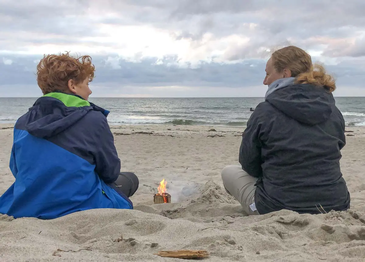 Mutter und Kind in Jacken sitzen an einem Sandstrand mit Blick auf das Meer, zwischen ihnen brennt ein kleines Feuer. Der Himmel ist bewölkt und die Atmosphäre wirkt ruhig und entspannt.