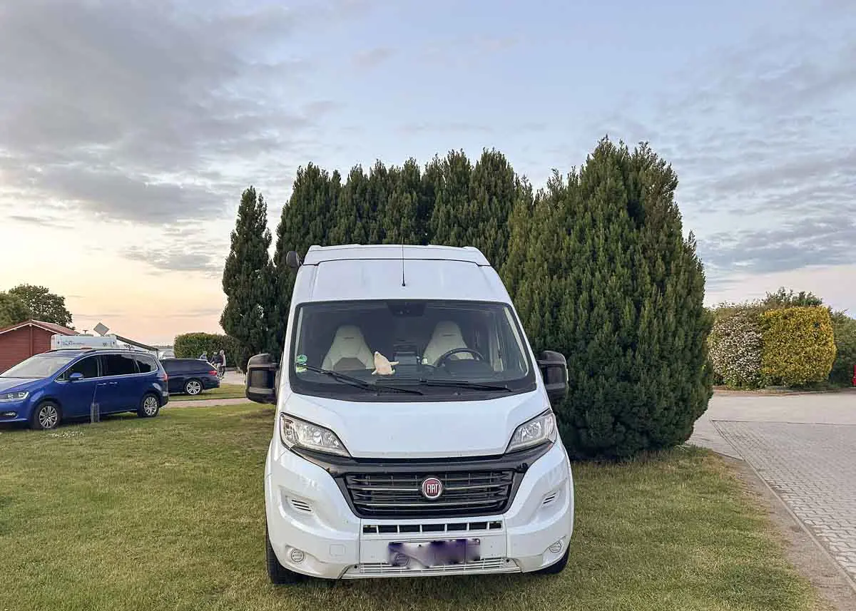 Ein weißes Wohnmobil steht auf einer Wiese vor hohen grünen Bäumen auf einem der landschaftlich reizvollen Ostsee-Campingplätze. Im Hintergrund sind andere Fahrzeuge und ein rotes Gebäude zu sehen, der Himmel ist teilweise bewölkt.