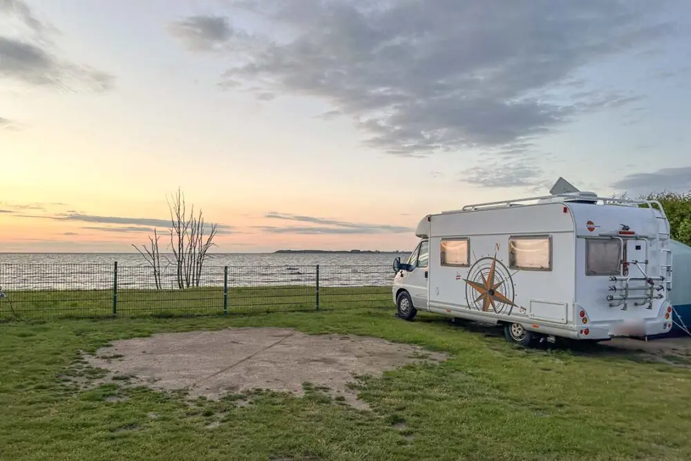 Ein weißes Wohnmobil steht bei Sonnenuntergang auf einer Wiese neben einem Metallzaun auf einem Ostsee Campingplatz am Strand und blickt auf das ruhige Meer.