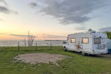 Ein weißes Wohnmobil steht bei Sonnenuntergang auf einer Wiese neben einem Metallzaun auf einem Ostsee Campingplatz am Strand und blickt auf das ruhige Meer.