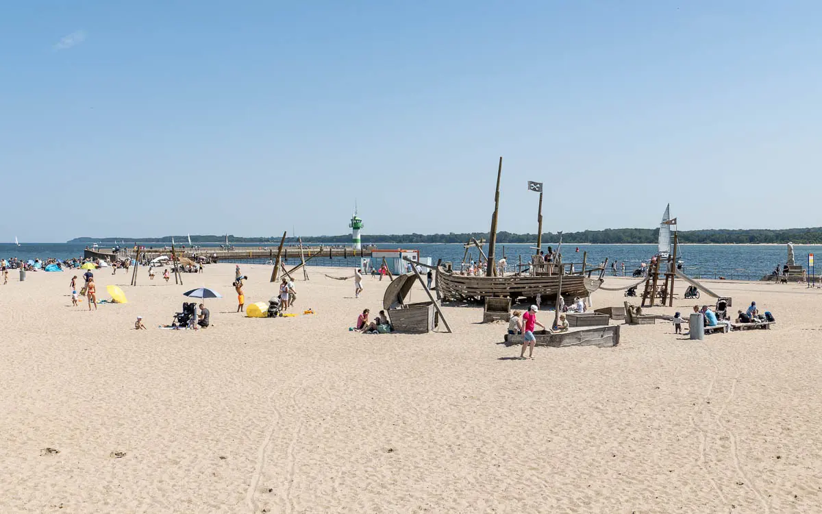 Die Menschen entspannen und spielen am Sandstrand in der Nähe des Wassers. Mit einem Piraten-Spielplatz genießen Familien Ostsee Camping. Ein grüner Leuchtturm steht in der Ferne unter einem klaren blauen Himmel.