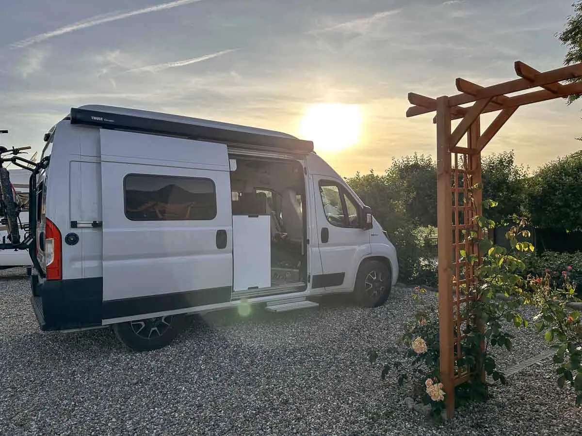 Ein weißes Wohnmobil mit offener Seitentür steht bei Sonnenuntergang auf einem Wohnmobilstellplatz aus Schotter. Rechts ist ein Holzspalier mit blühenden Blumen zu sehen, umgeben von üppigen Büschen im Hintergrund.