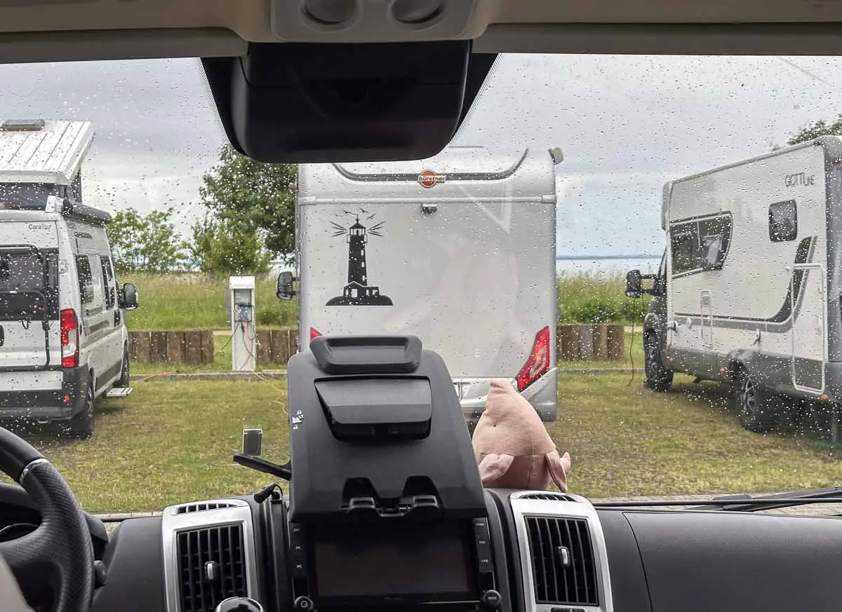 Blick aus dem Inneren eines Wohnmobils durch eine verregnete Windschutzscheibe auf drei geparkte Wohnmobile auf einem Wohnmobil-Stellplatz, mit Bäumen und bewölktem Himmel und im Hintergrund die Ostsee. Im Vordergrund sind ein Armaturenbrett und ein kleines Spielzeugschwein zu sehen.