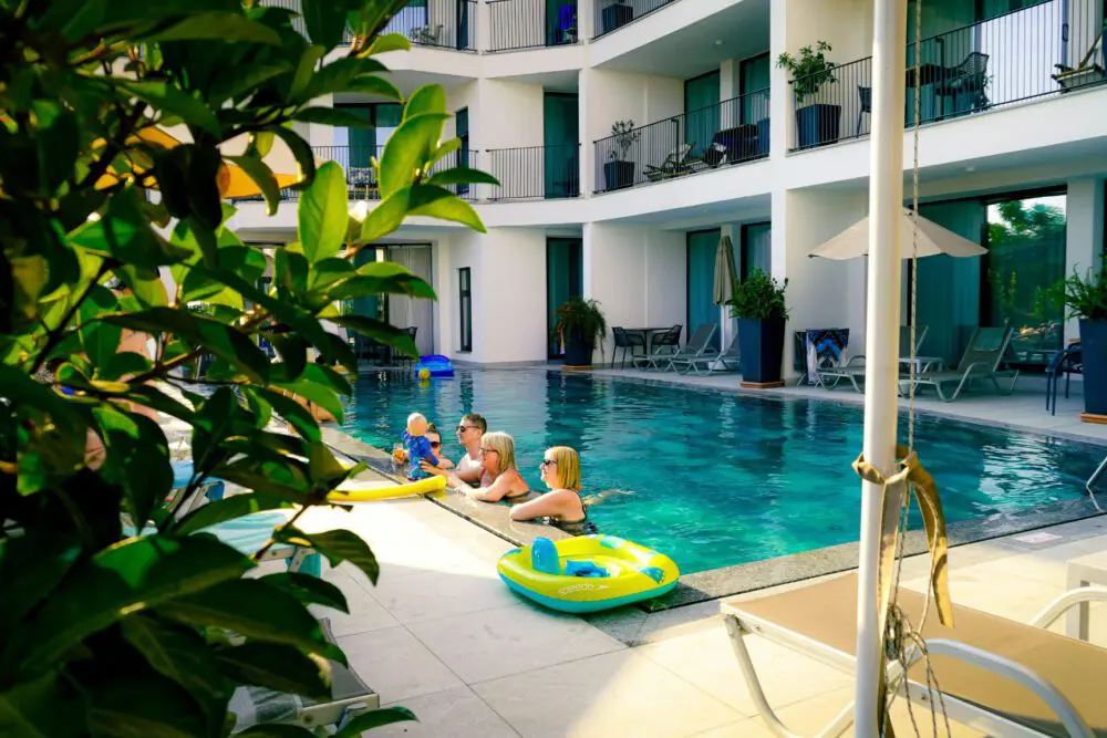 Mehrere Kinder spielen auf aufblasbaren Spielzeugen in einem familienfreundlichen Hotelpool, umgeben von modernen Balkonen und Liegestühlen, mit grünen Pflanzen im Vordergrund und Sonnenlicht, das die Szene hervorhebt.