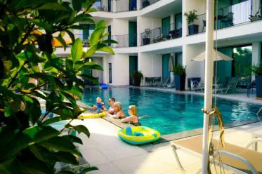 Mehrere Kinder spielen auf aufblasbaren Spielzeugen in einem familienfreundlichen Hotelpool, umgeben von modernen Balkonen und Liegestühlen, mit grünen Pflanzen im Vordergrund und Sonnenlicht, das die Szene hervorhebt.