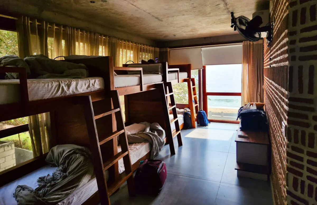 Ein Herbergszimmer mit vier hölzernen Etagenbetten, unordentlichen Laken und Koffern wird von einem großen Fenster mit Blick auf das Meer erhellt, mit Vorhängen auf der einen Seite und einem Ventilator über einer Bank, auf der sich Taschen stapeln - so sieht ein kinderfreundliches Hotel definitiv nicht aus.