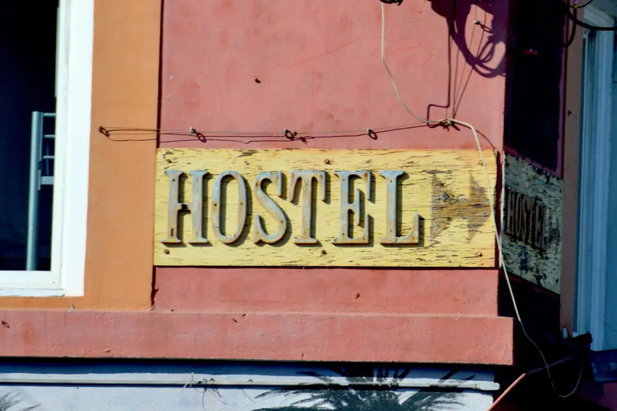 Auf einem verwitterten gelben Schild mit großen, erhabenen Buchstaben steht HOSTEL an der rosa und orange gestrichenen Außenwand einer familienfreundlichen Unterkunft. Einige verblasste Farbe und sichtbare Drähte umgeben das Schild.