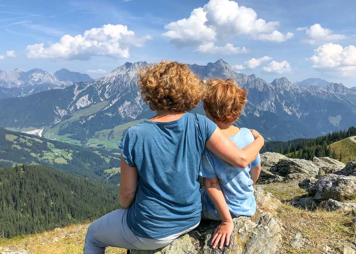 Zwei Menschen mit lockigem Haar, von hinten gesehen, sitzen auf einem Felsvorsprung mit Blick auf eine malerische Berglandschaft mit grünen Tälern, Wäldern und einem blauen, wolkenverhangenen Himmel. Eine Person hat ihren Arm um die andere gelegt.