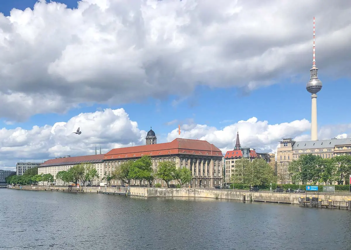 BERLIN-Fernsehturm-Spree-5948 Ein Blick auf Berlin vom Flussufer aus mit historischen Gebäuden, grünen Bäumen und dem berühmten Fernsehturm unter einem teilweise bewölkten Himmel. Ein Vogel fliegt in der Nähe der Gebäude.