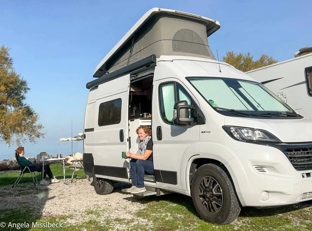 Ein weißer Wohnwagen steht an einem See. Eine Person sitzt in der offenen Seitentür und hält eine Tasse in der Hand, während eine andere Person draußen an einem Tisch sitzt. Im Hintergrund sind Bäume und Wasser zu sehen.