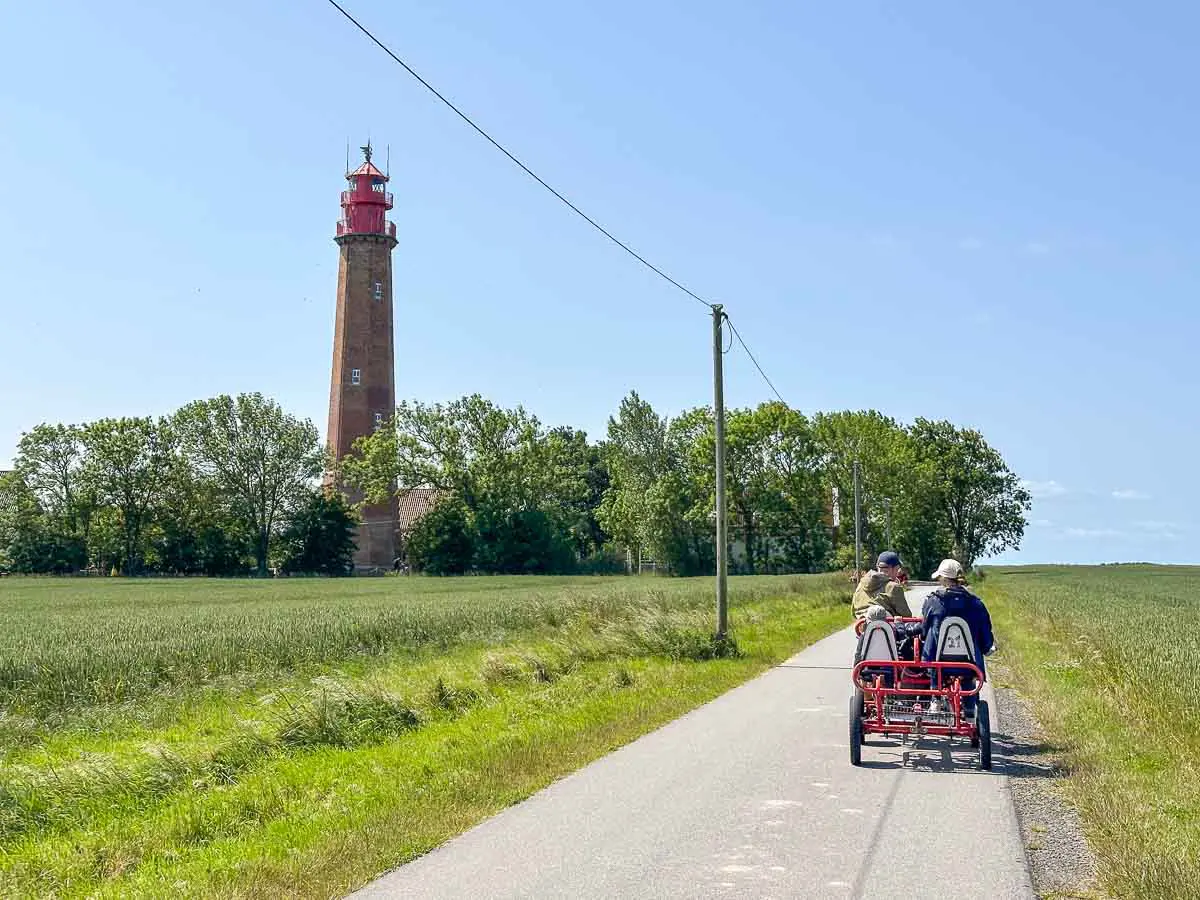 Eine Familie fährt mit einem vierrädrigen Fahrrad eine von Gras gesäumte Landstraße entlang, im Hintergrund sind der hohe rote Leuchtturm Flügge und Bäume vor einem strahlend blauen Himmel zu sehen.