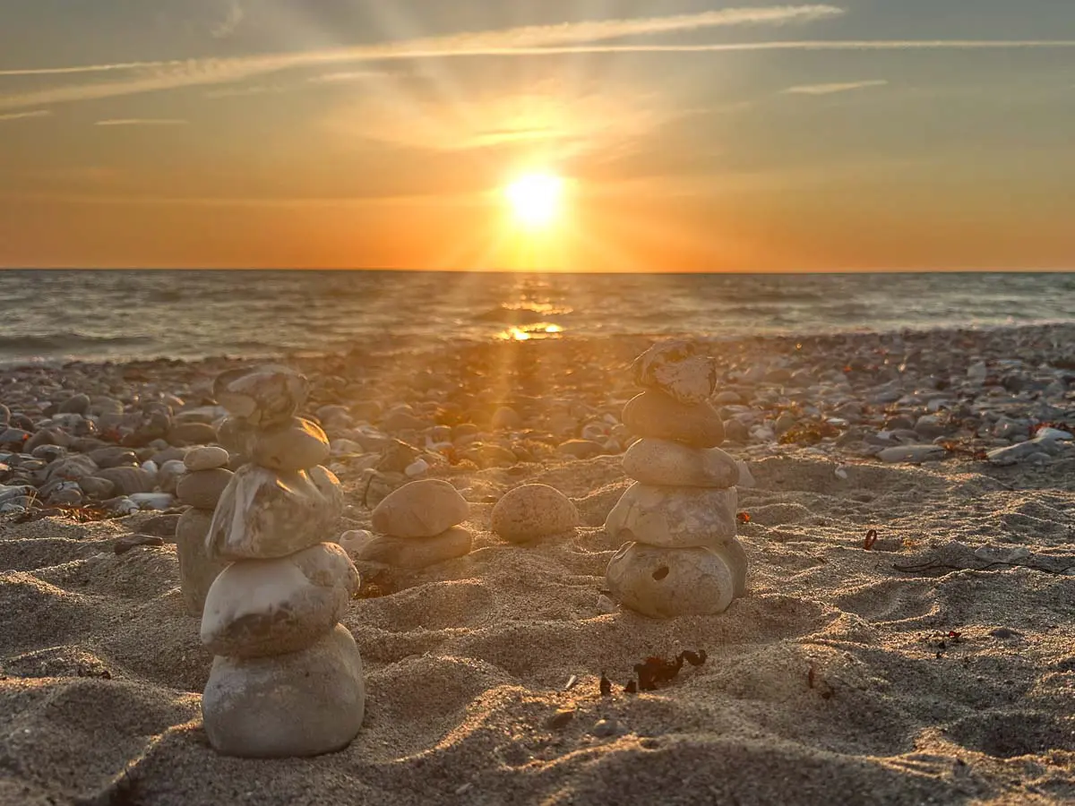 Sonnenuntergang über einem ruhigen Meer, im Vordergrund aufgeschichtete Steinmännchen an einem Sandstrand; Sonnenstrahlen breiten sich am Himmel aus und tauchen den ganzen Flügger Strand auf Fehmarn in einen warmen, goldenen Schein.