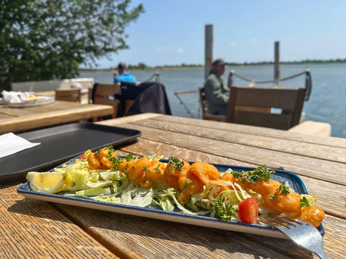 Auf einem Holztisch in der Aalkate im Freien am Wasser in Fehmarn steht ein Teller mit Garnelensalat, garniert mit Kräutern und Zitrone, während sich im Hintergrund an einem sonnigen Tag Menschen entspannen.