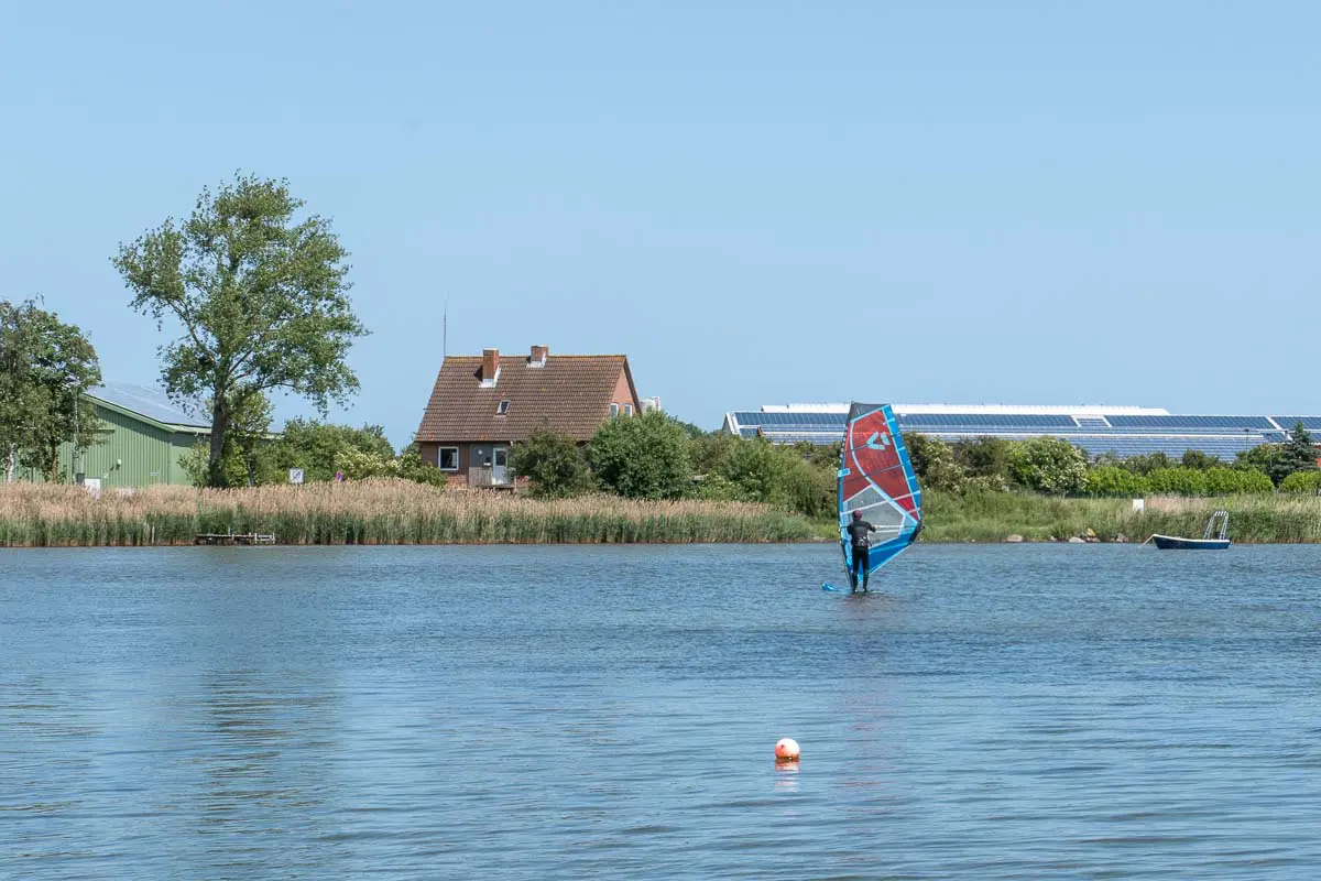 Eine Person, die auf  ruhigem Meer in der Nähe vom Goldstrand auf Fehmarn windsurft, im Hintergrund ein Haus, Grünflächen und Solaranlagen unter einem strahlend blauen Himmel. Im Vordergrund schwimmt eine Boje im Wasser.