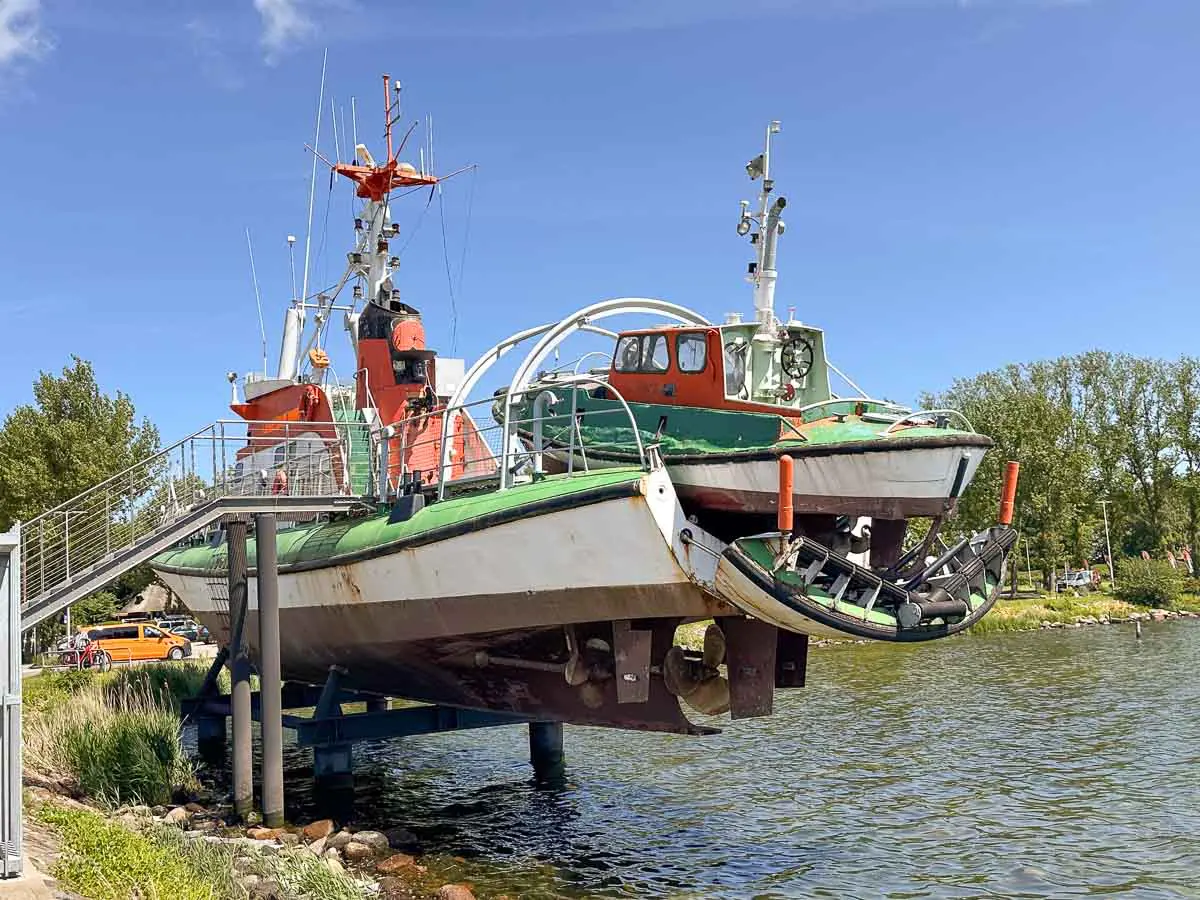 Ein grün-weißes Rettungsboot mit Ausliegerboot ist auf einer Metallrampe über dem Wasser ausgestelt, zu der eine Treppe führt. Im Hintergrund sind Bäume und der blaue Himmel zu sehen. So bietet der Hafen von Burgstaaken ein Abenteuer auf Fehmarn mit Kindern“. 