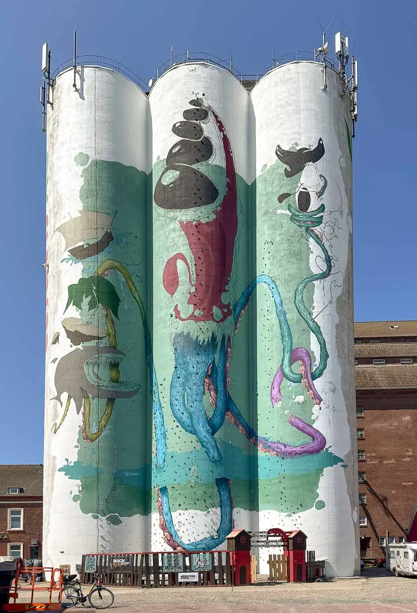 Ein großes Wandgemälde mit einem farbenfrohen Tintenfisch und abstrakten Formen ziert drei hohe, zylindrische Silos, die zum Klettern auf Fehmarn mit Kindern einladen vor dem Hintergrund eines strahlend blauen Himmels und alter Hafengebäude.