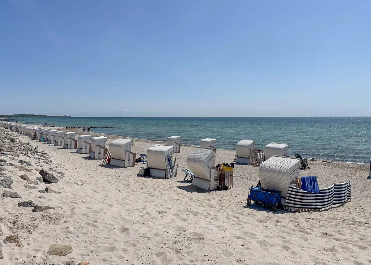 Weiße Strandkörbe aus Korbgeflecht mit Sonnendächern säumen den Sandstrand auf Fehmarn mit Kindern und blicken unter einem strahlend blauen Himmel auf das ruhige Meer. Ein paar Menschen entspannen an diesem friedlichen, familienfreundlichen Strand in Burgtiefe.
