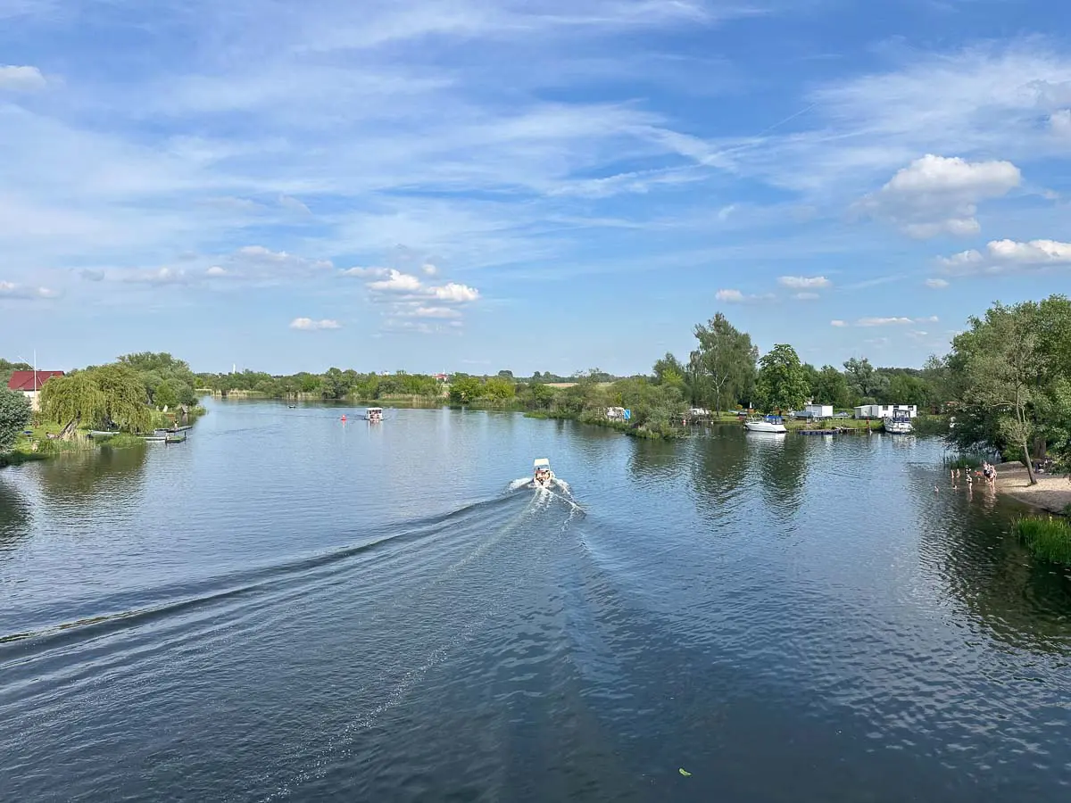 Ein kleines Motorboot fährt auf der Havel zwischen Brandenburg und Rathenow im Westhavelland, umgeben von grünen Bäumen und grasbewachsenen Ufern unter einem strahlend blauen Himmel mit vereinzelten Wolken. Am rechten Ufer sind Menschen zu sehen.