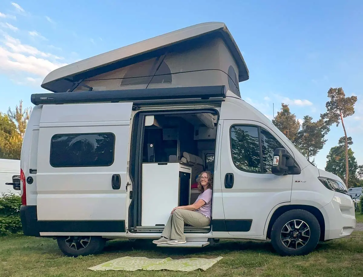 Eine Person sitzt in der offenen Seitentür eines weißen Wohnmobils mit Aufstelldach, das auf einer Wiese auf einem Campingplatz im malerischen Havelland geparkt ist, mit Bäumen und blauem Himmel im Hintergrund. Vor dem Wohnmobil liegt eine grün gemusterte Matte.