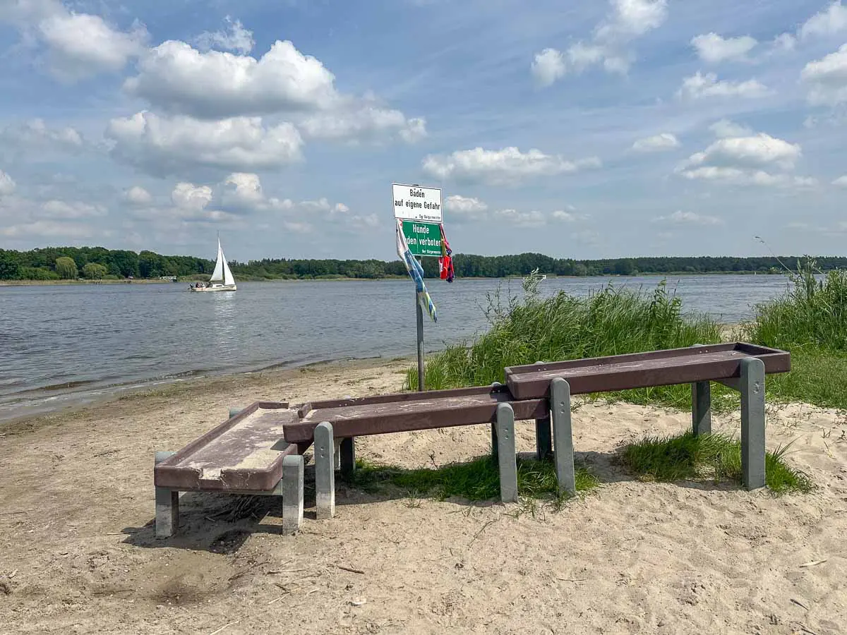 An einem sandigen Strand am Seeufer im Havelland sind im Vordergrund eine Wasserspielanlage aus Holz, ein Wegweiser, eine Rasenfläche und ein kleines Segelboot zu sehen, das unter einem teilweise bewölkten Himmel über das Wasser gleitet.