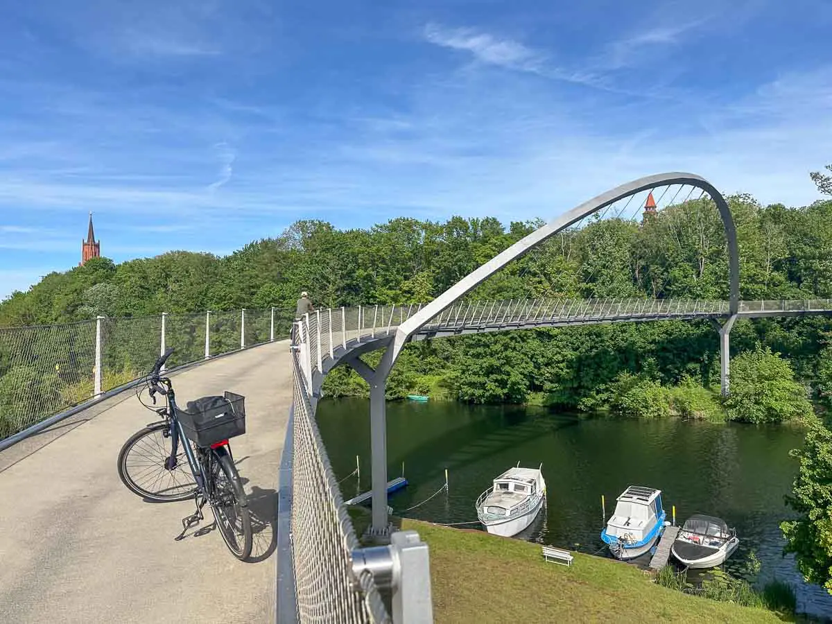 Eine moderne Fußgängerbrücke mit einem geschwungenen Bogen überspannt die Havel in Rathenow. Darunter liegen Boote vor Anker. Auf der Brücke steht ein Fahrrad, und im Hintergrund ragen unter einem strahlend blauen Himmel Bäume und ein Kirchturm empor.
