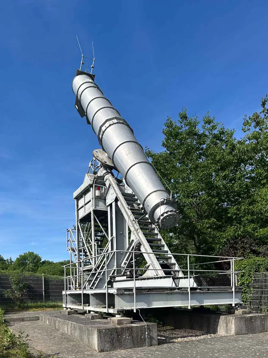Ein großes, silbernes Teleskop im Industriestil ist im Freien in Rathenow im Havelland auf einer Metallplattform montiert und in Richtung Himmel ausgerichtet, im Hintergrund sind Grünflächen und ein blauer Himmel zu sehen. Ein Metallzaun umgibt das Gelände.