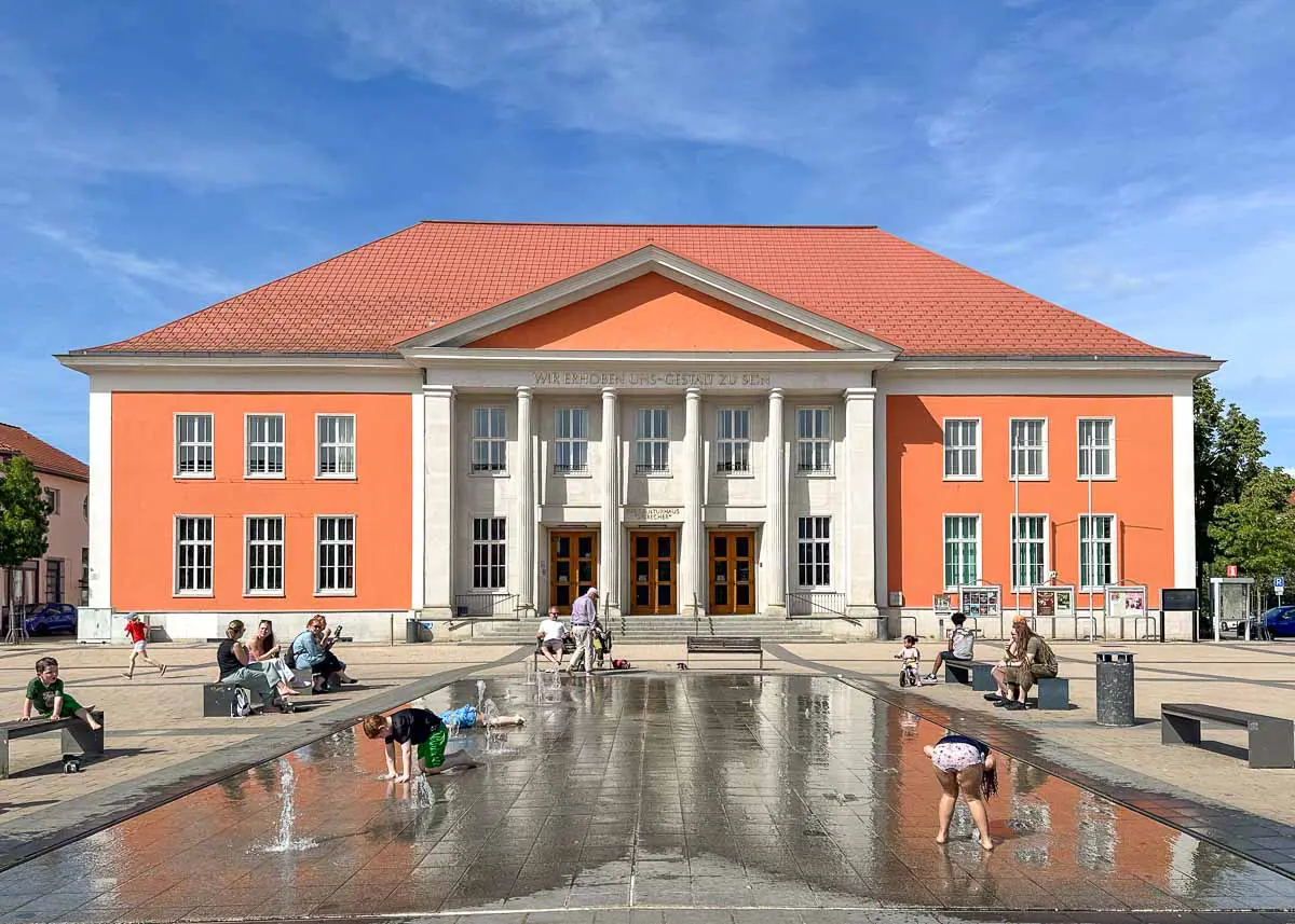 In Rathenow im Havelland spielen Kinder in einem ebenerdigen Springbrunnen vor dem Optikmuseum, einem großen, orange-weißen Gebäude im neoklassizistischen Stil mit rotem Dach, während Erwachsene in der Nähe auf Bänken unter dem blauen Himmel sitzen.