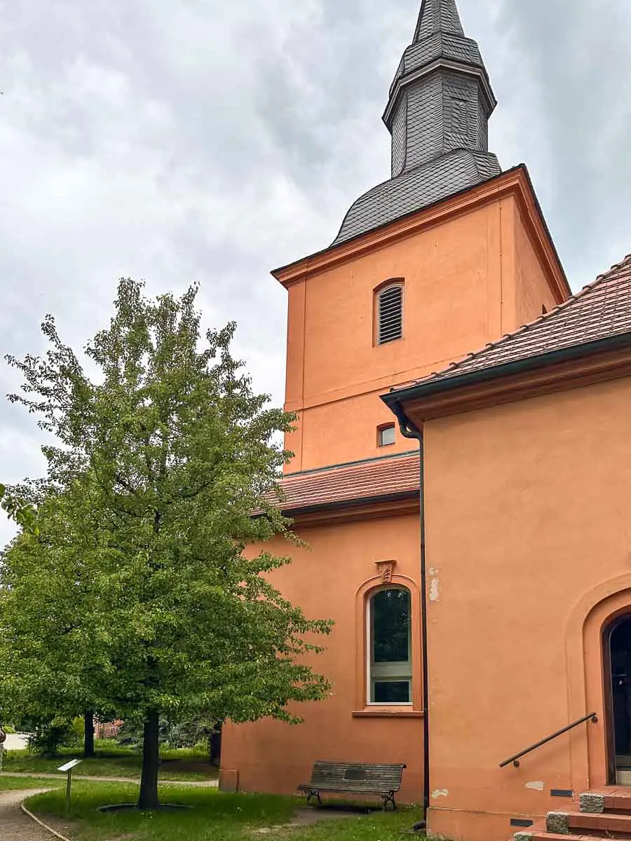 Im Herzen von Havelland steht eine orange-pfirsichfarbene Kirche mit einem hohen Kirchturm neben einem üppig grünen Baum. In der Nähe des Eingangs steht eine Bank, während sich über ihr graue Wolken zusammenziehen und der Szene eine ruhige Atmosphäre verleihen.