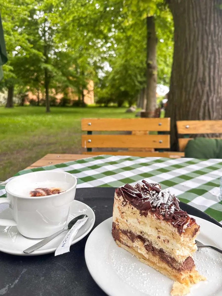 Ein Stück Schichtkuchen mit Schokolade und Puderzucker sowie eine Tasse Cappuccino stehen auf einem Tisch mit grün kariertem Tischtuch im Freien in Havelland, mit Blick auf einen Park mit Bäumen und Holzbänken.