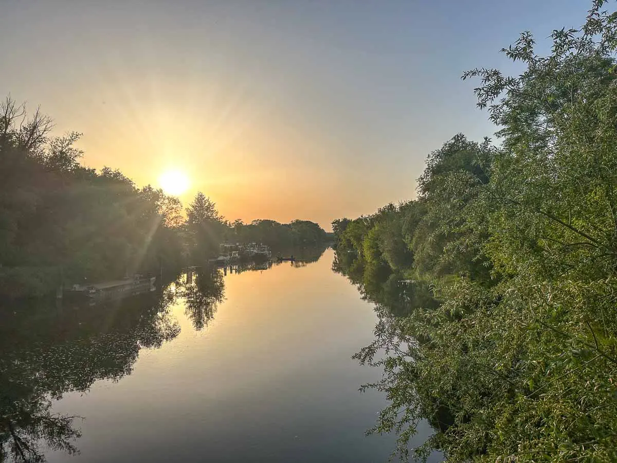 Die Sonne geht über einem ruhigen, von Bäumen gesäumten Fluss im Havelland unter, taucht die Landschaft in ein warmes, goldenes Licht und lässt den Himmel und das Laubwerk im stillen Wasser spiegeln. Dichte, grüne Bäume säumen beide Ufer dieser friedlichen Szenerie.