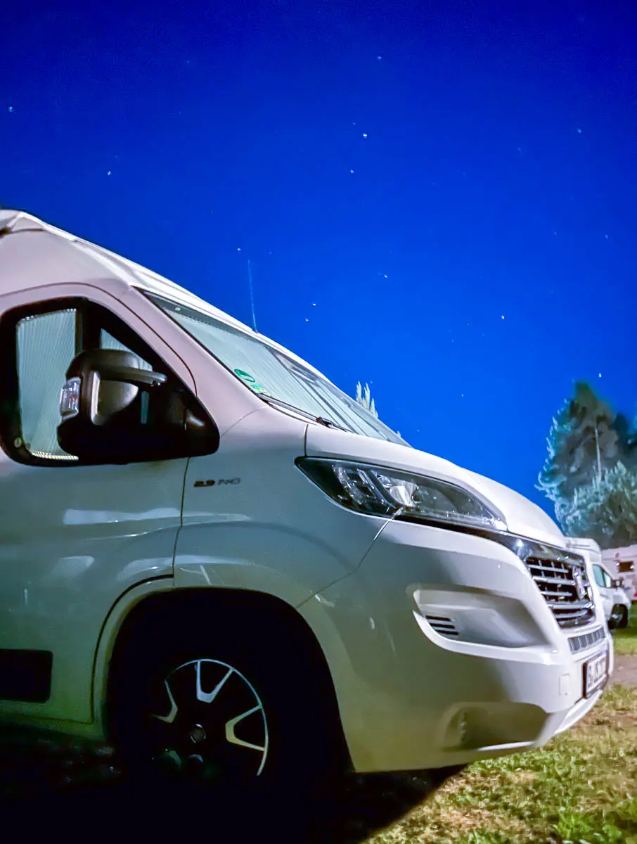 Ein weißer Wohnmobil steht unter dem klaren, sternenklaren Nachthimmel des Havellands, im Hintergrund sind Bäume und Campingausrüstung zu sehen. Das Fahrzeug wird von vorne links betrachtet.