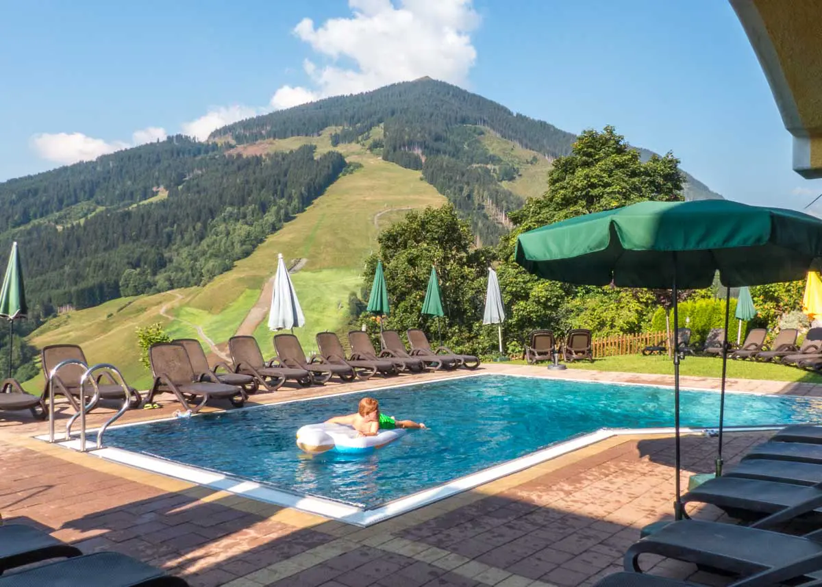 Ein Kind treibt auf einem aufblasbaren Schwimmring in einem klaren Außenpool, der von Liegestühlen und Sonnenschirmen umgeben ist, vor dem Hintergrund grüner Berge und eines blauen Himmels.