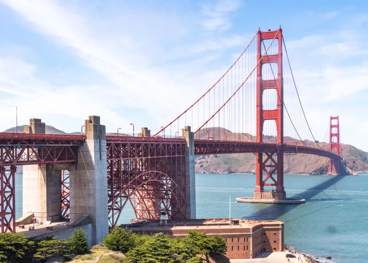 WELTWEIT-Golden-Gate-Bridge-San-Francisco-1090464 An einem sonnigen Tag in San Francisco erstreckt sich die Golden Gate Bridge über das blaue Wasser, im Hintergrund sind Hügel zu sehen, und am Fuße der Brücke liegt Fort Point.