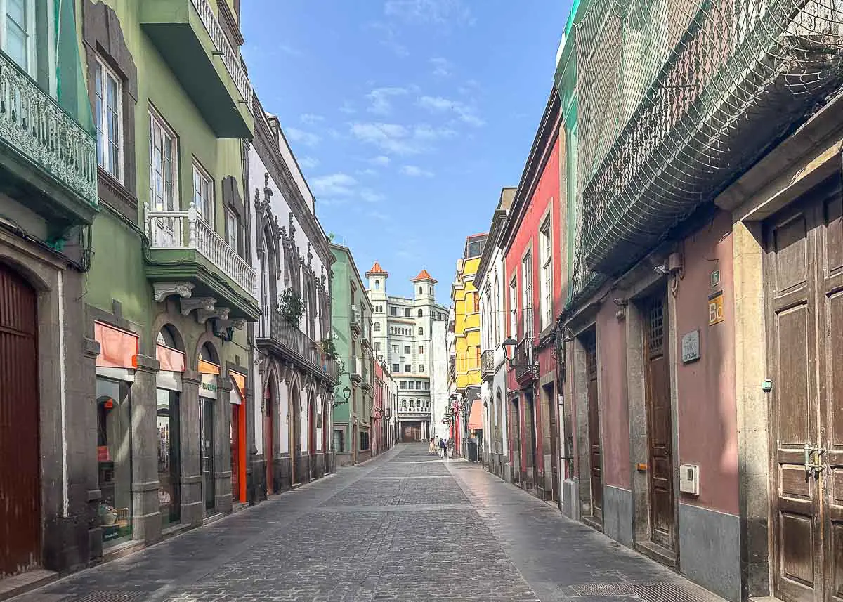 Eine schmale Kopfsteinpflasterstraße in der Altstadt Vegueta, gesäumt von farbenfrohen Gebäuden mit Balkonen und kunstvollen Details, erstreckt sich unter einem blauen Himmel mit leichten Wolken bis in die Ferne – eine der bezaubernden Sehenswürdigkeiten in Las Palmas de Gran Canaria.